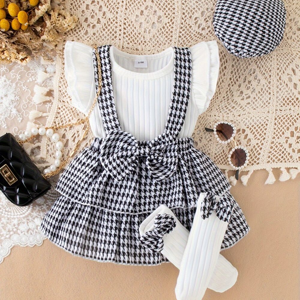 Toddler girl houndstooth suspender skirt set with ribbed white top, ruffled sleeves, pleated front, and black & white houndstooth skirt with waist bow, displayed for catalog
