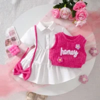 Girls white dress and pink knitted vest set with embroidery, floral appliqués, tiered skirt, and matching accessories, displayed for catalog