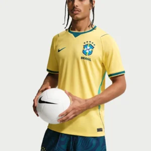 A man with braided hair modeling the official yellow 2026 Brazil national football team home jersey while holding a white football, as seen in the product